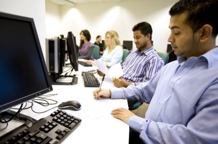 Male trainee in foreground, sitting in front of a monitor writing. Other trainees in the background doing the same.